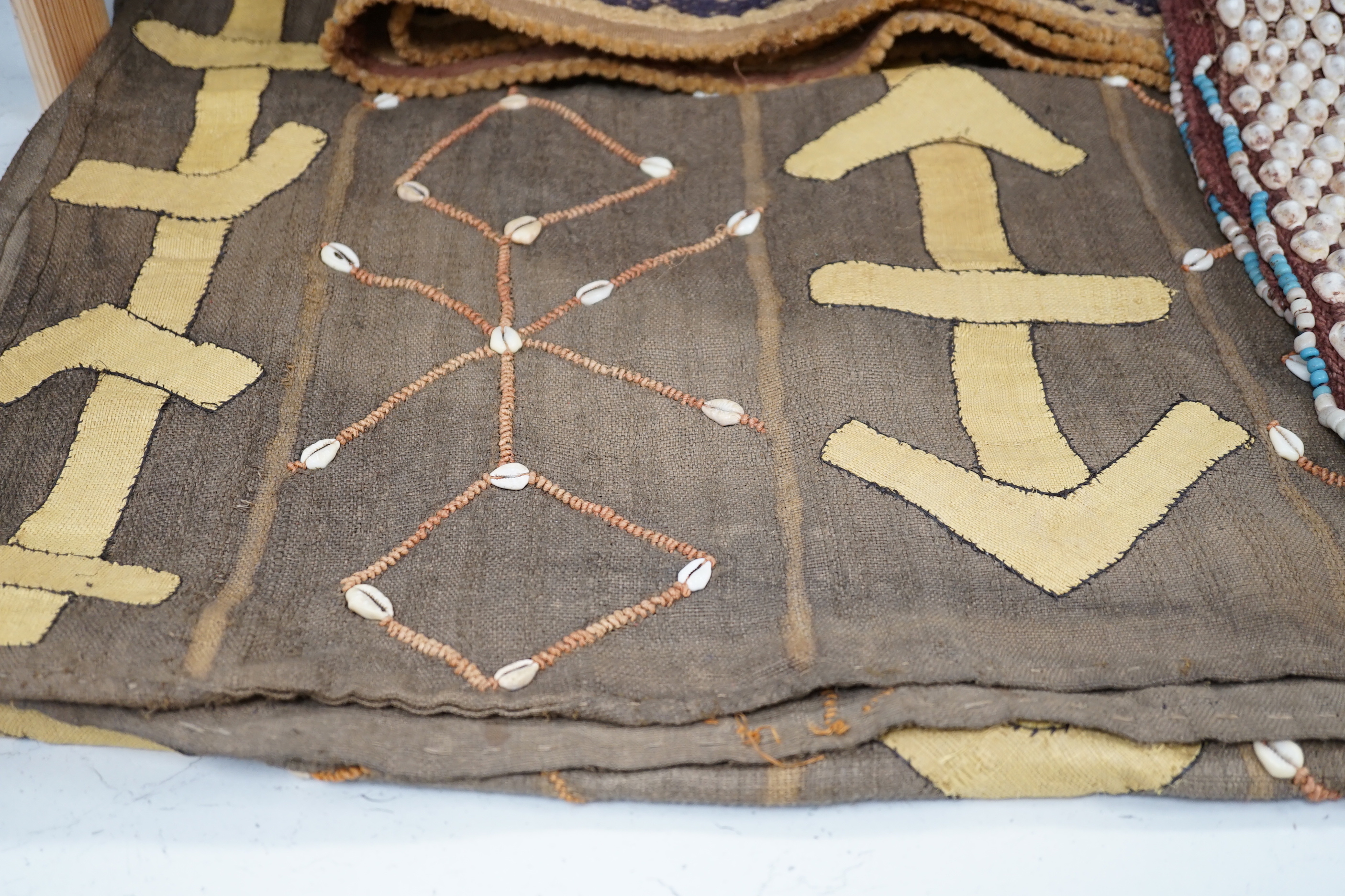 Three African tribal hangings, embroidered with coloured hand dyed threads and appliquéd shells on smaller handwoven cloth panels all sewn together to form the hangings, including a chieftains coloured bead worked and sh